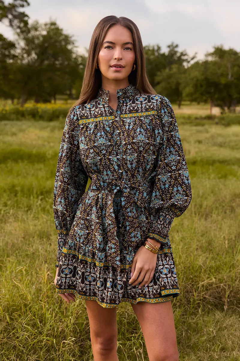 A woman stands in a grassy outdoor field wearing a long-sleeve mini dress with a detailed geometric and tribal-style pattern in brown, blue, black, and yellow.