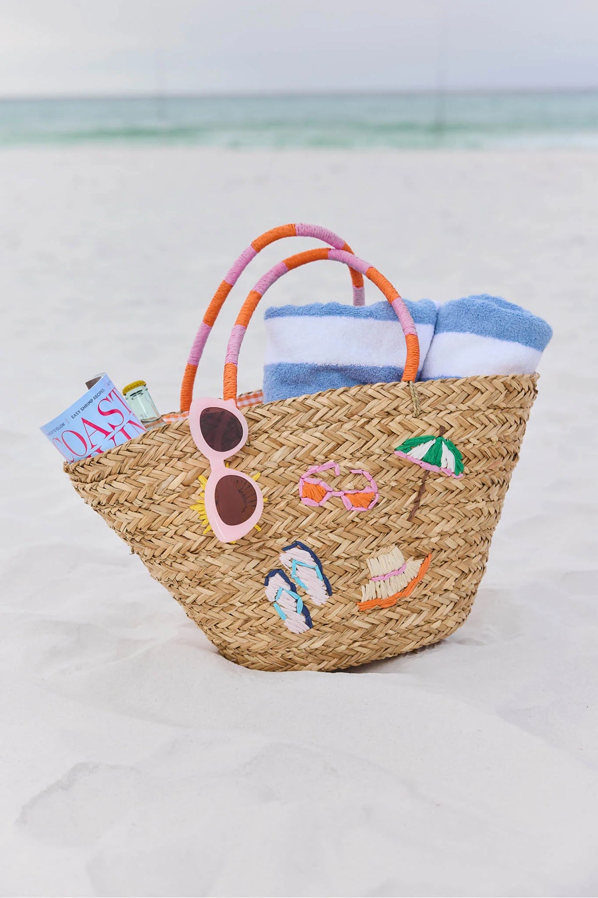 Natural woven raffia beach tote with colorful icon embroidery on sandy beach - Lifestyle Shot | Avara