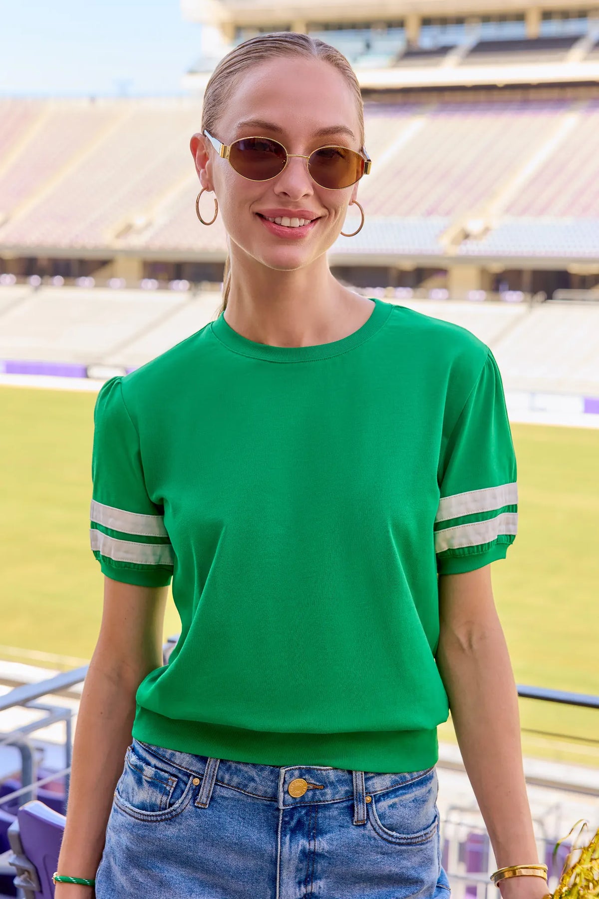 A woman wearing a bright green short-sleeve knit top with white stripes on the sleeves, paired with blue jeans and round sunglasses, standing in a stadium setting.
