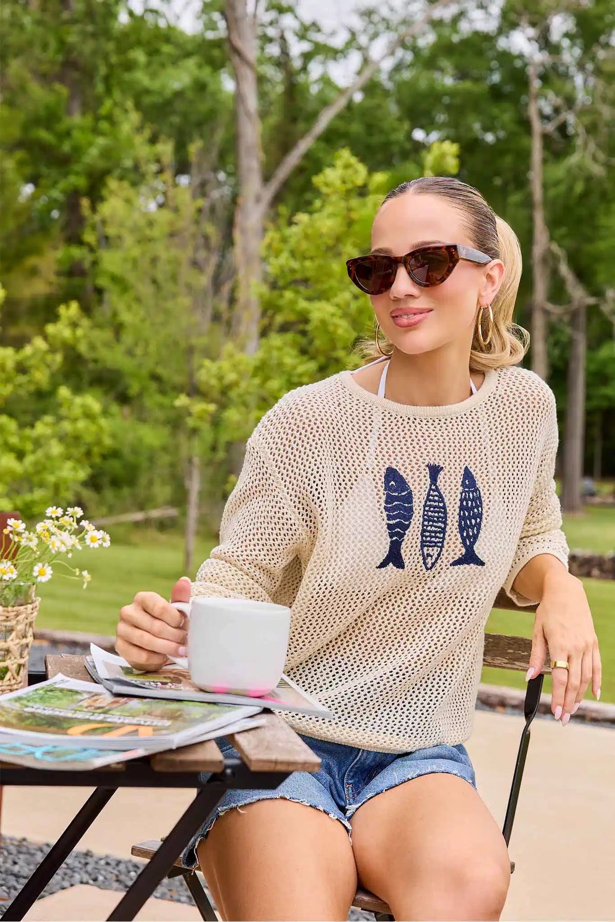 Woman sitting outdoors at a table with a scenic background, wearing sunglasses and a casual outfit.