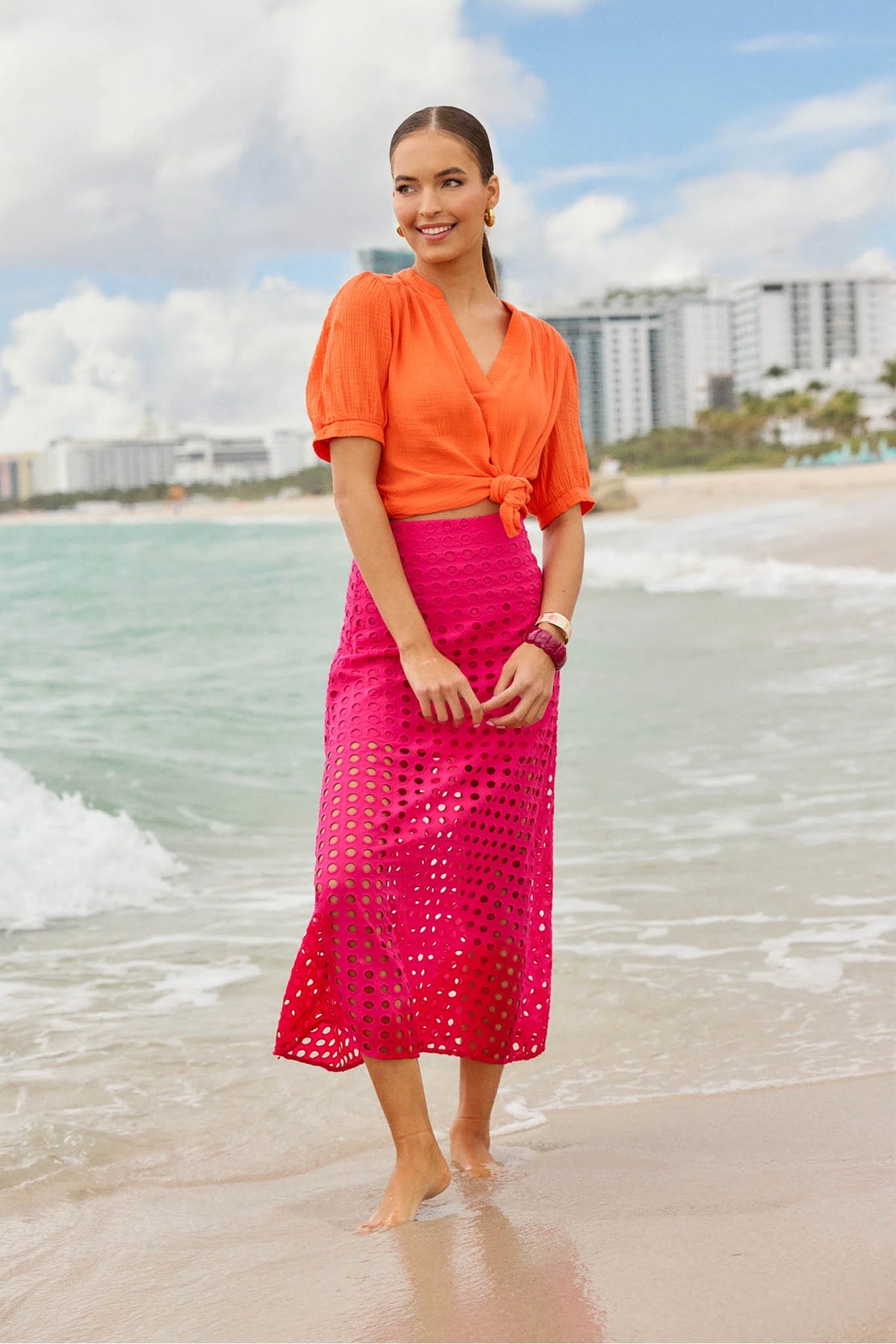 Woman at beach in Ellie Pink Eyelet Midi Skirt, featuring a midi A-line silhouette with eyelet embroidery, paired with a knotted blouse, smiling serenely.