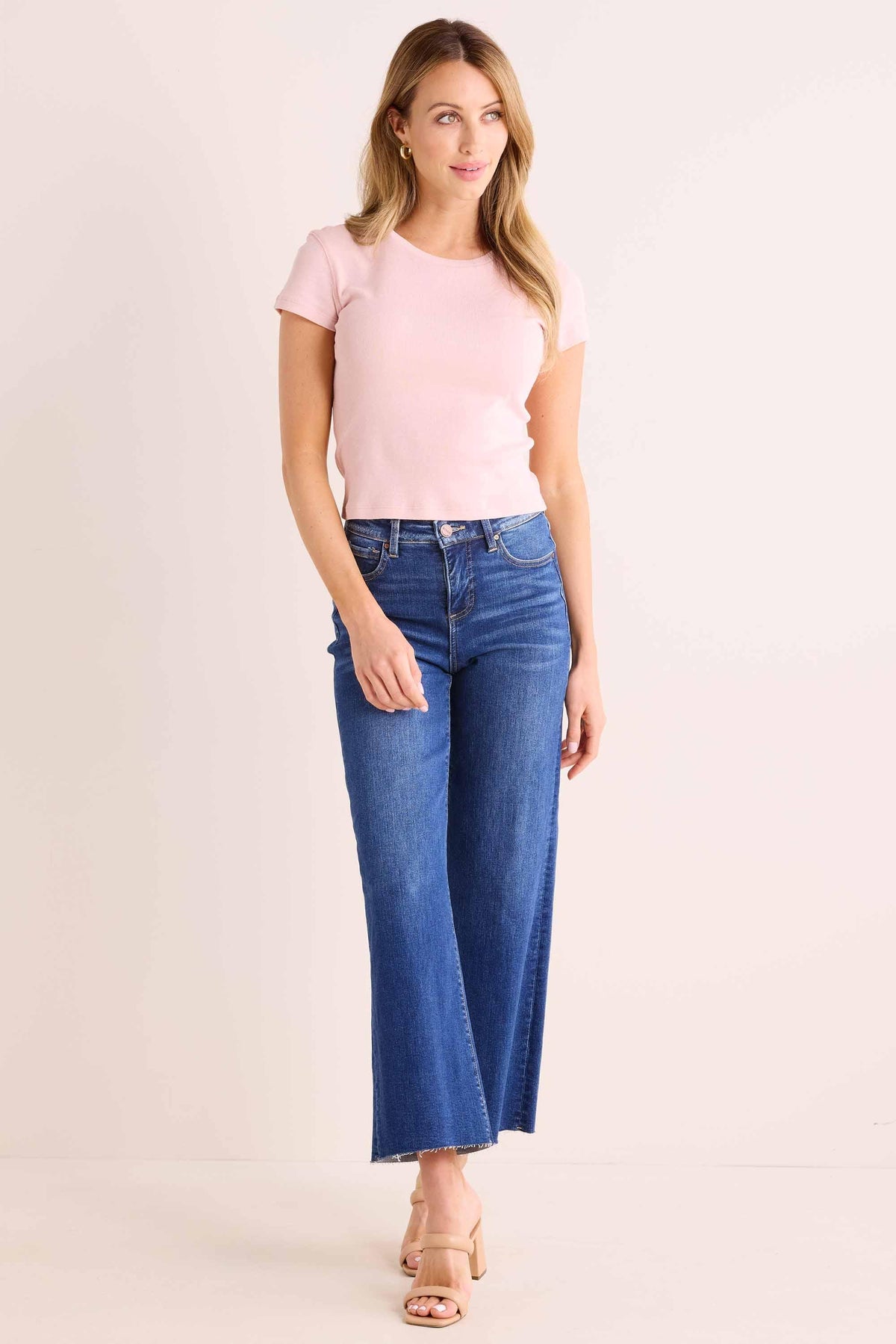 Woman models Aria Pink Fitted Tee, baby pink crew neck, short sleeves, styled with wide-leg jeans and tan heeled sandals | Avara