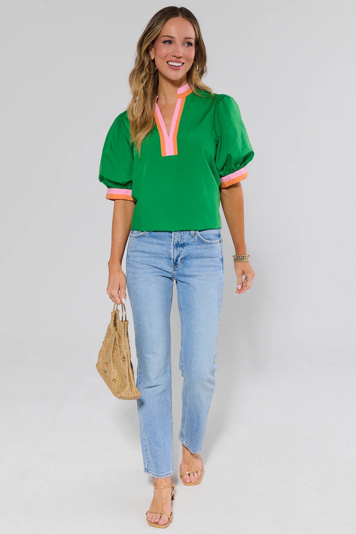 Woman wearing a green top with pink accents and blue jeans, holding a beige handbag on a gray background