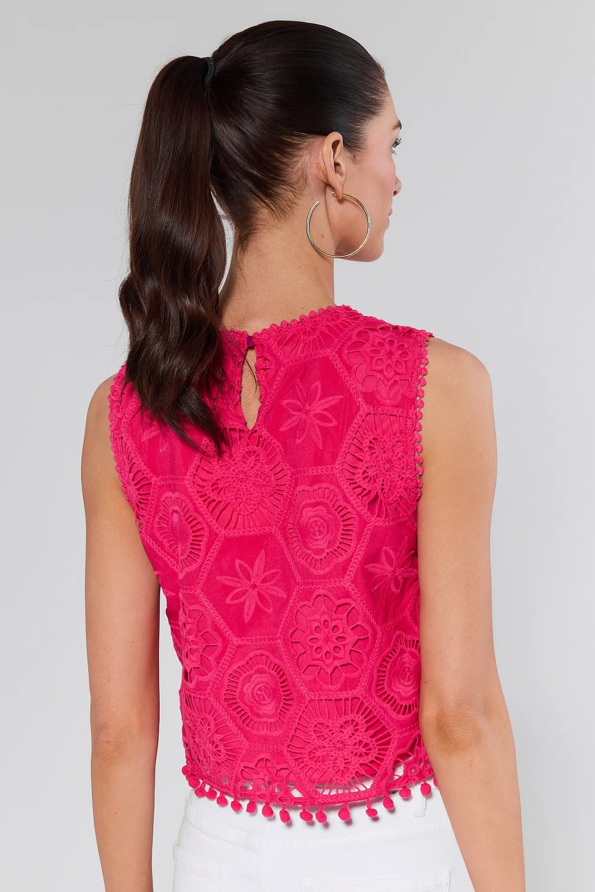 Back view of woman in Arden Pink Eyelet Top, featuring sleeveless design, hexagonal lace motifs, keyhole back, and pom-pom trim, paired with white bottoms.