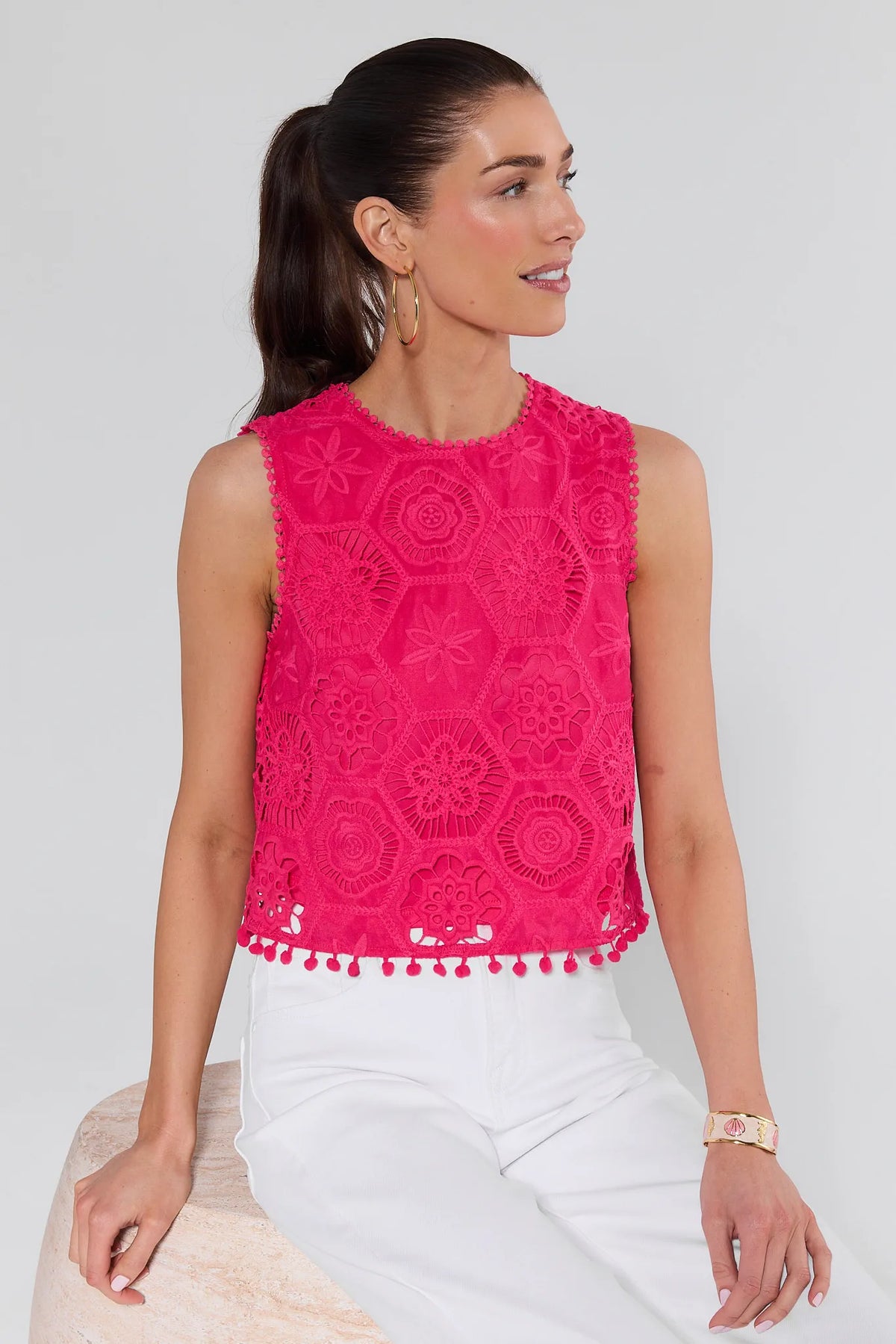 Woman wearing Arden Pink Eyelet Top with hexagonal floral lace, pom-pom hem, paired with high-waisted white pants, gold hoop earrings, and enamel cuff.