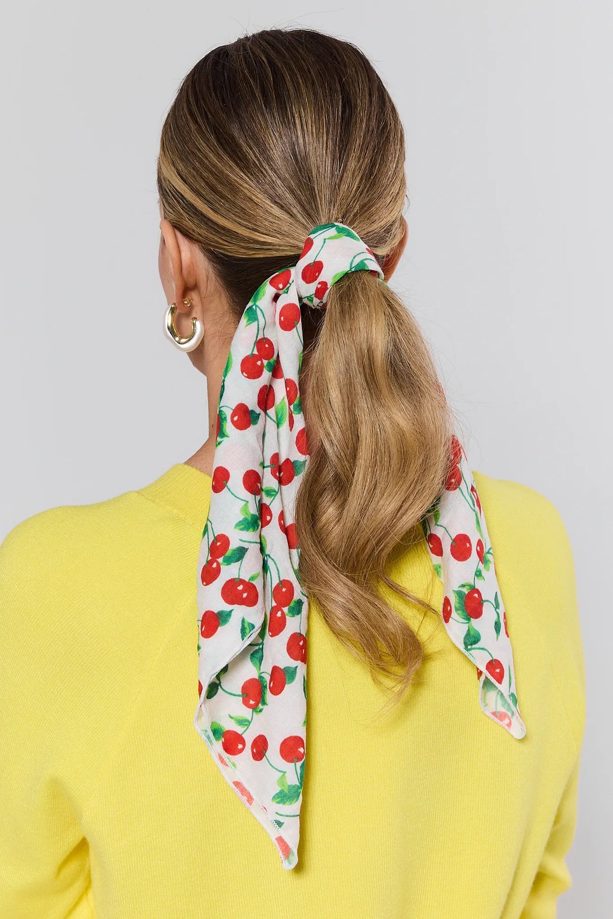 Person with a ponytail wearing a cherry-patterned scarf on a plain background