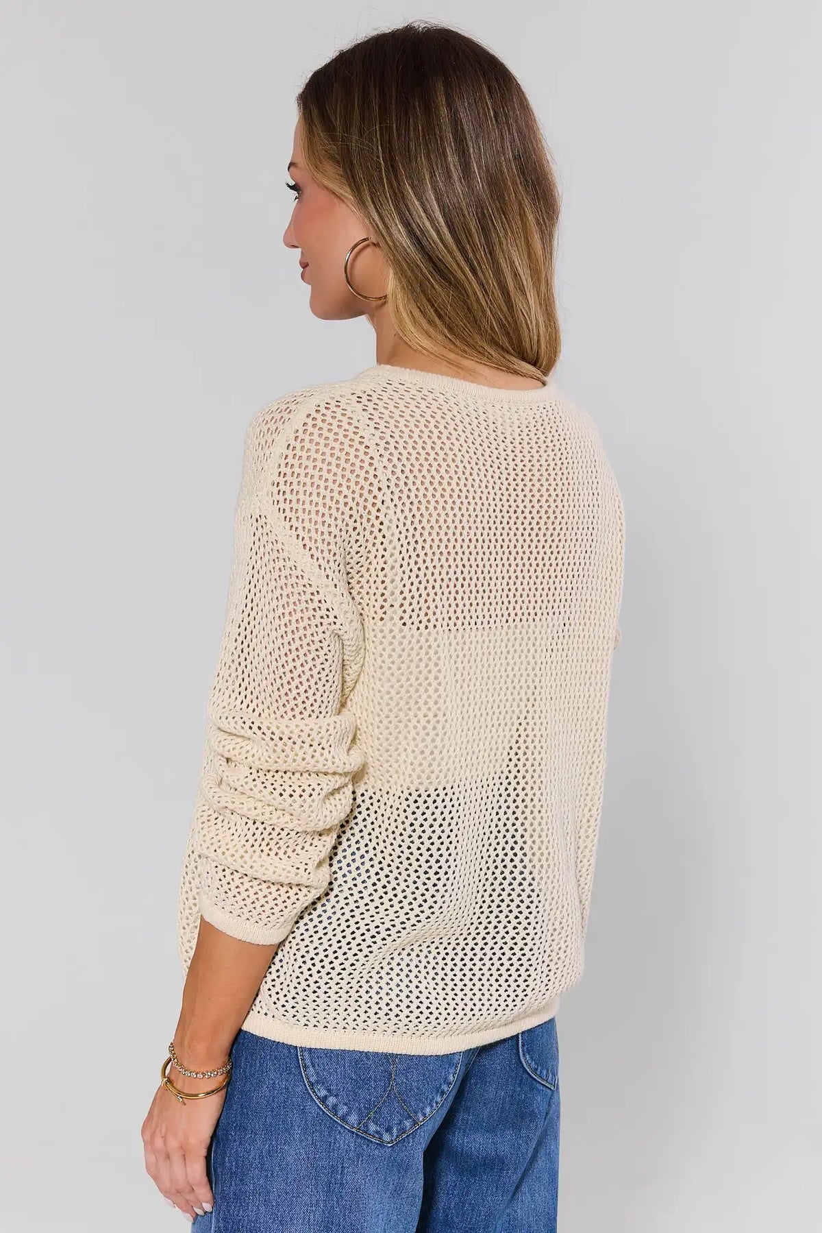 Woman standing wearing a beige knitted sweater and blue jeans against a plain background