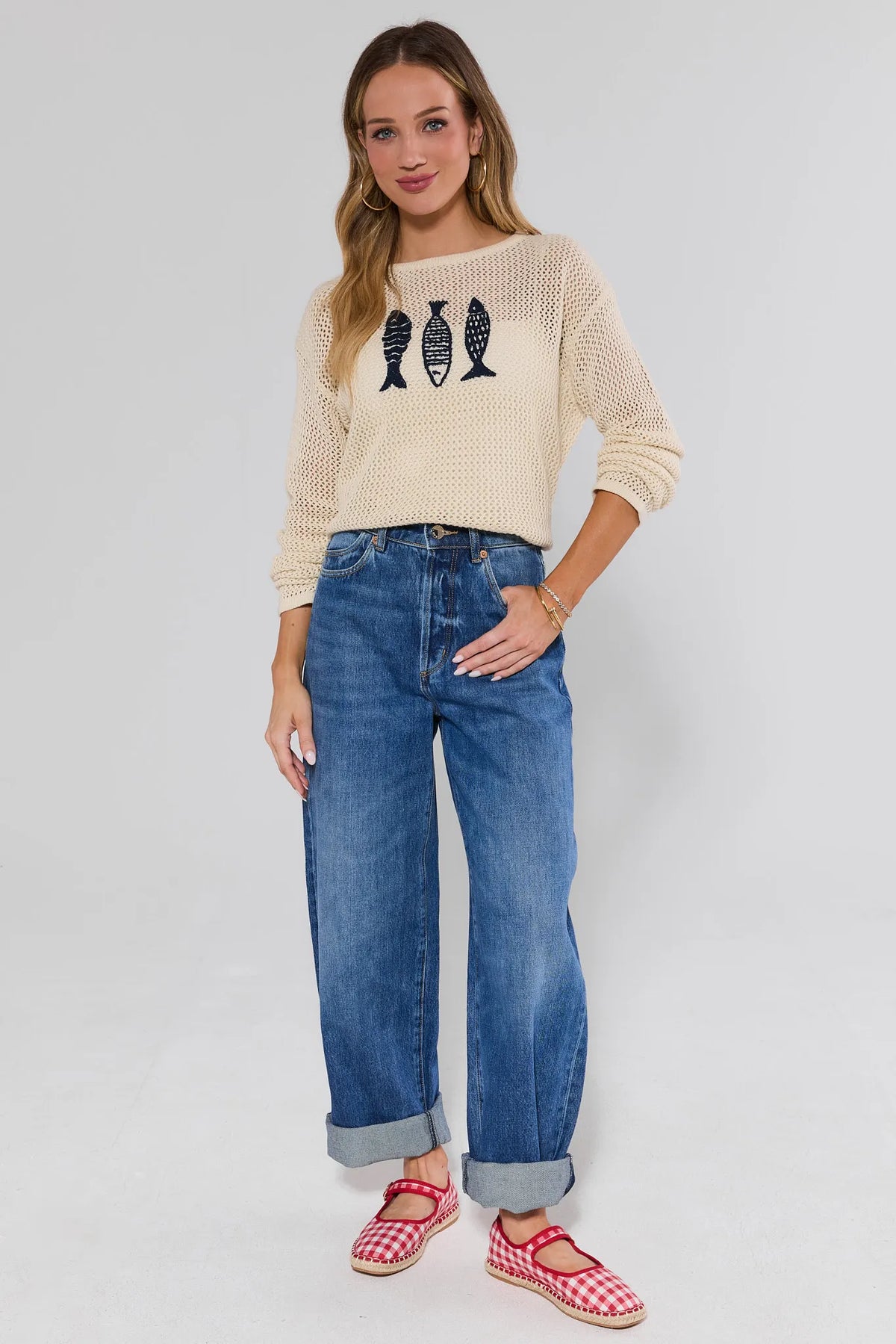 Woman wearing a beige sweater with fish design and blue jeans and gingham shoes on a white background