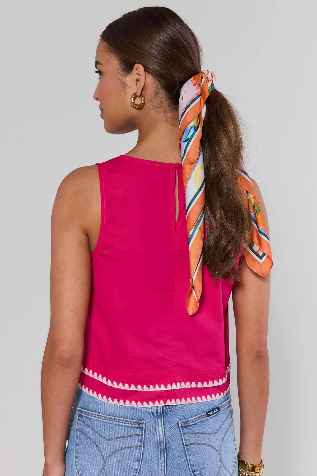 Woman wearing a pink top with colorful accents and blue jeans, standing against a plain background.