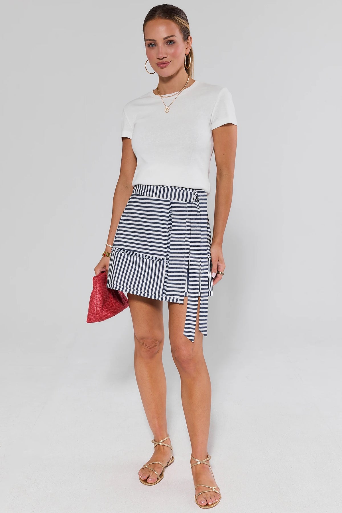 Woman wearing a white top and striped skirt with a red clutch against a white background