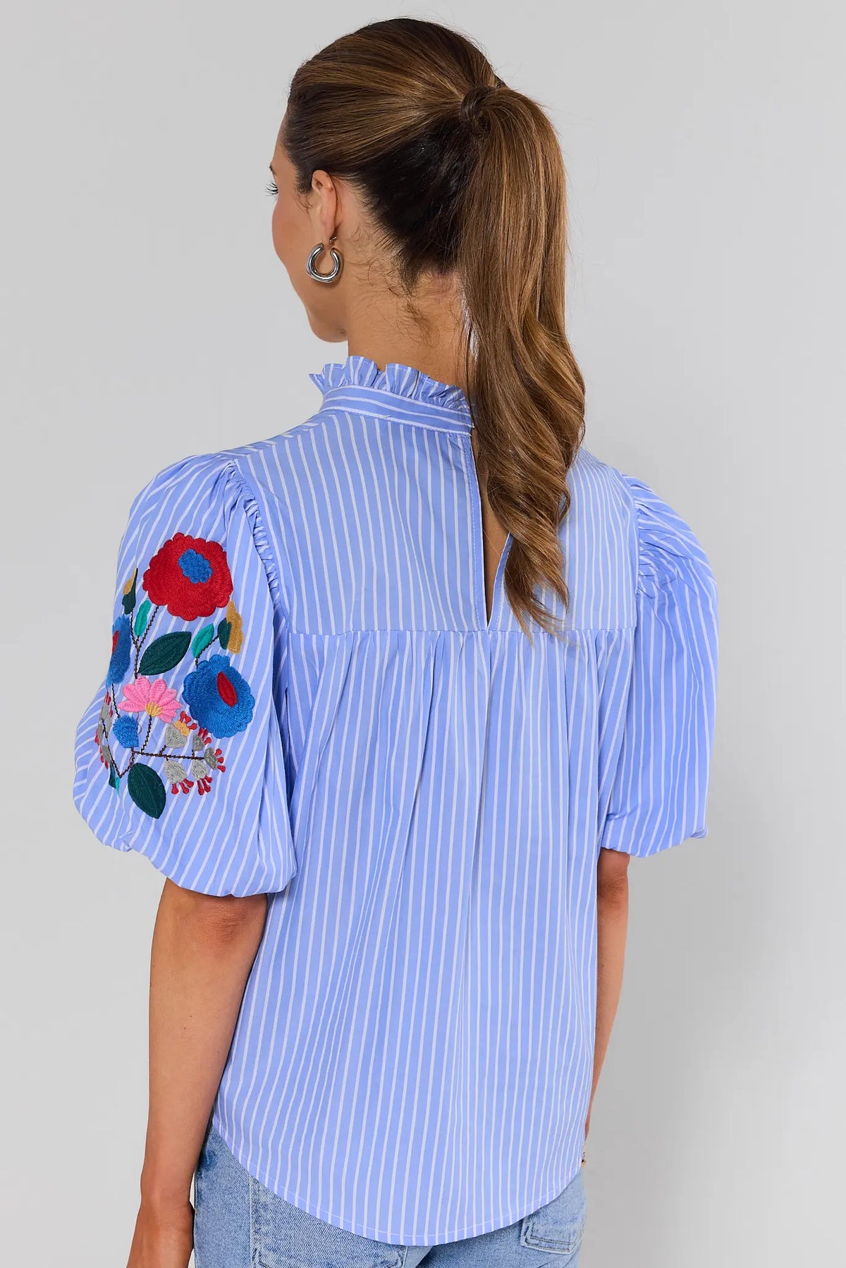 Woman standing wearing a blue striped blouse with floral embroidery on the sleeves against a gray background