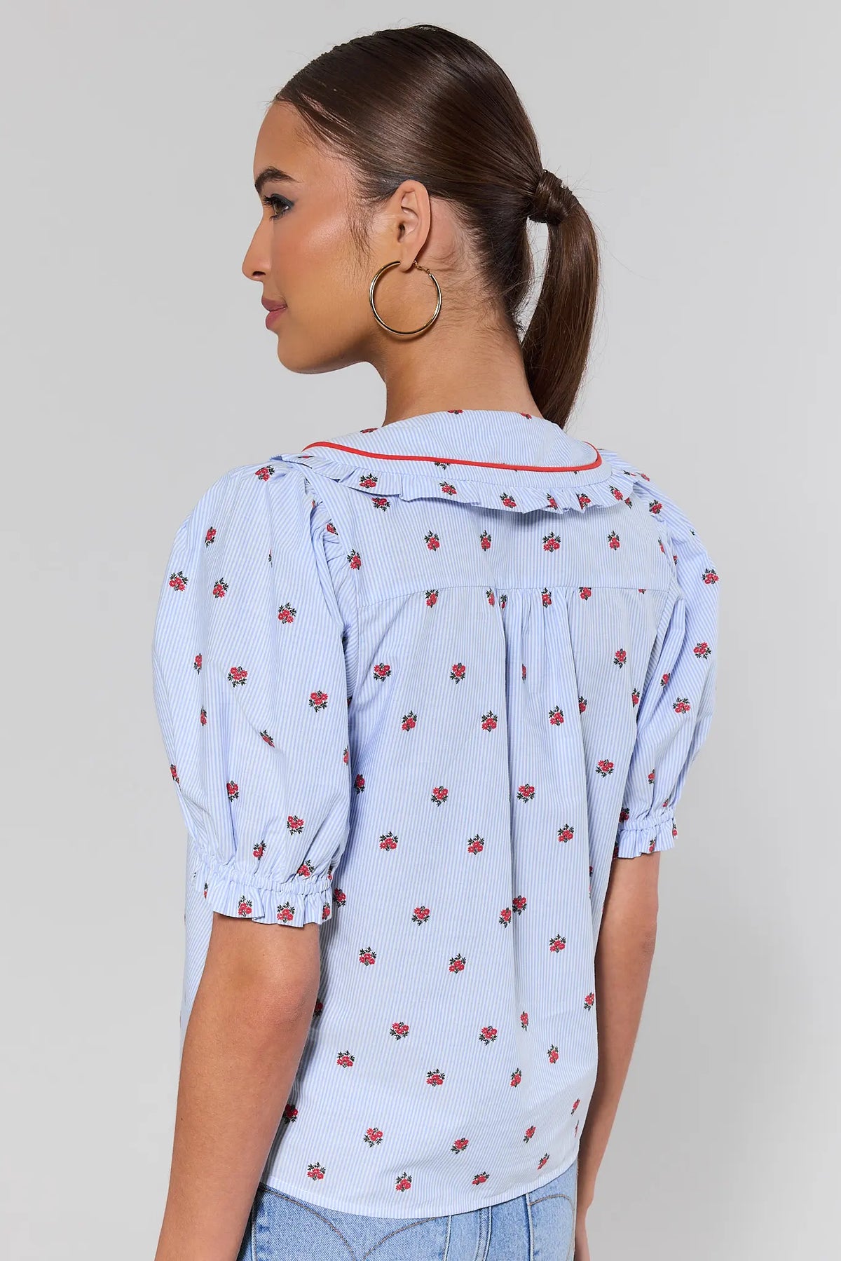 Woman standing wearing a light blue blouse with red floral pattern against a plain background