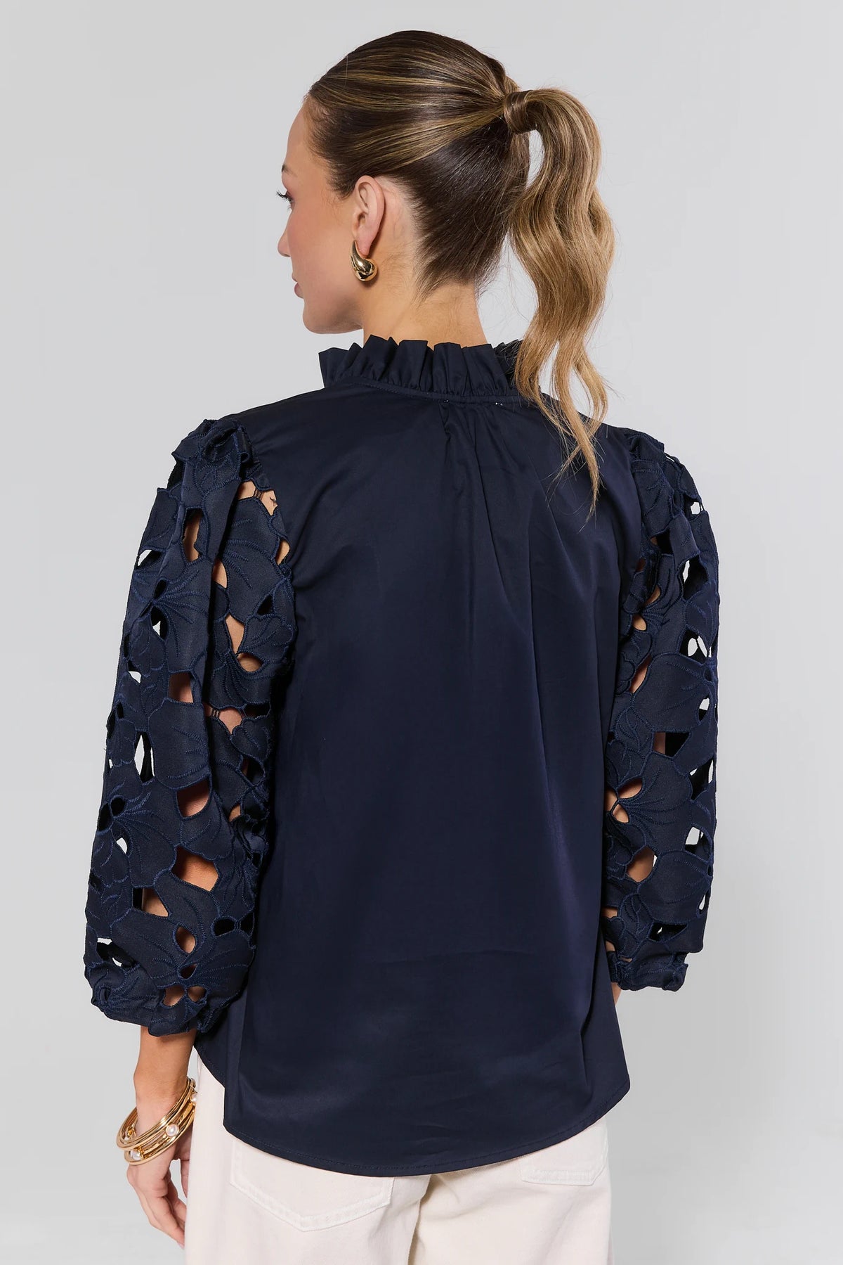 Back view of navy eyelet sleeve top by THML showing cutout lace sleeves and ruffled mock neckline, worn with gold accessories | Avara