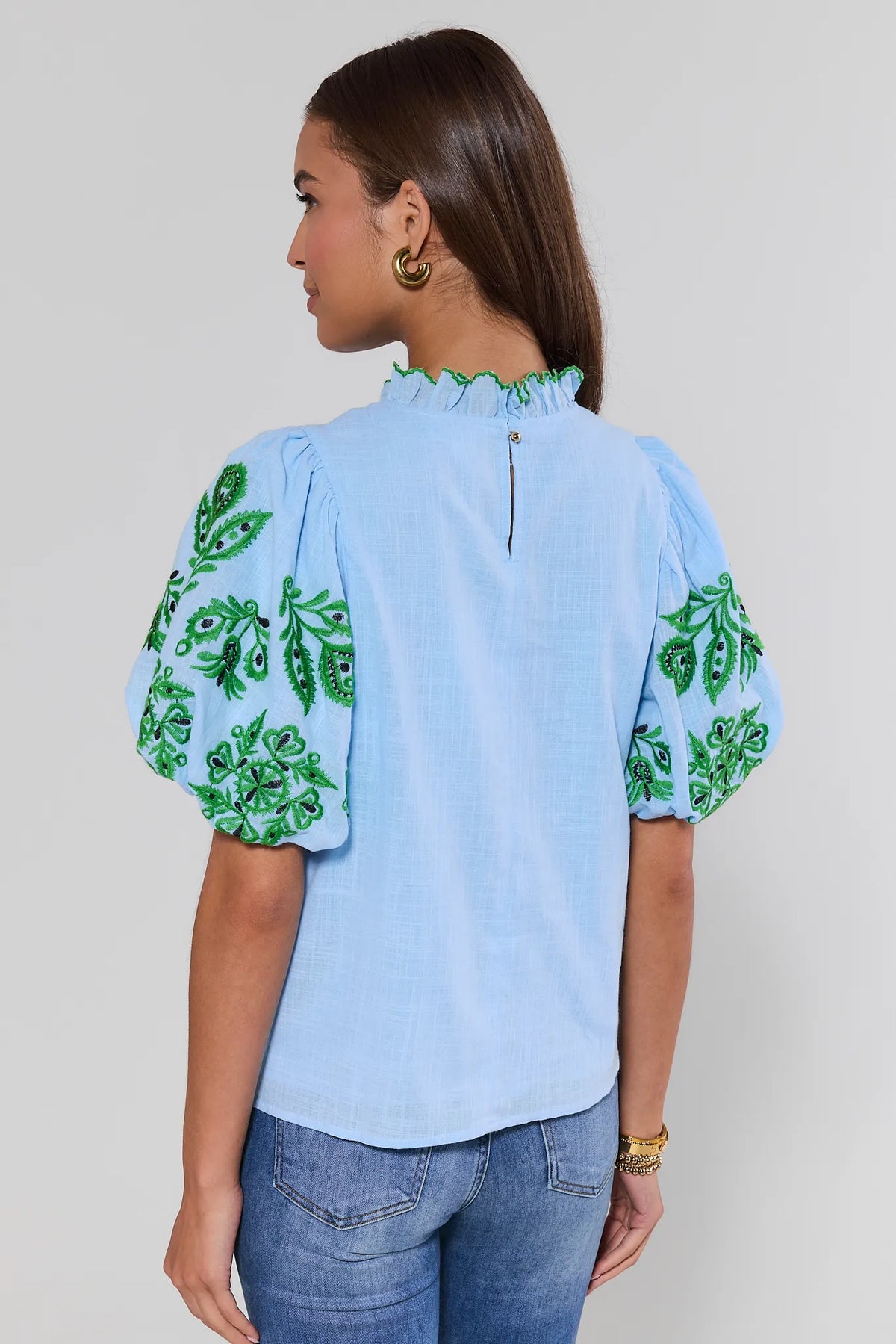 Woman standing wearing a light blue blouse with green floral patterns on a plain background