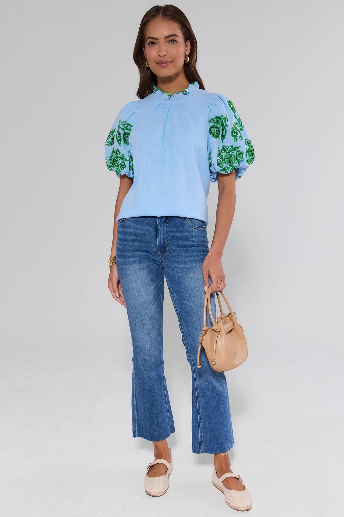 Woman wearing a light blue blouse with green floral patterns and blue jeans, holding a beige handbag on a white background.