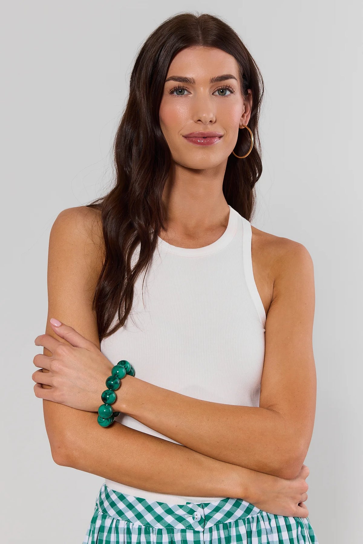 Woman wearing a white tank top and green checkered shorts with a green bracelet on a plain background