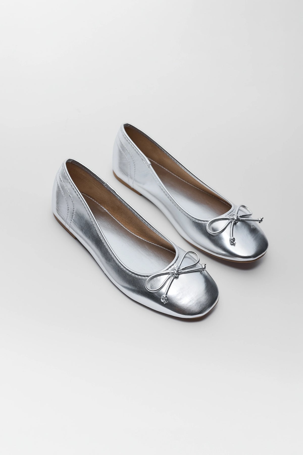 Metallic silver bow ballet flats pair, angled overhead view showing feminine bow detail on both shoes | Avara