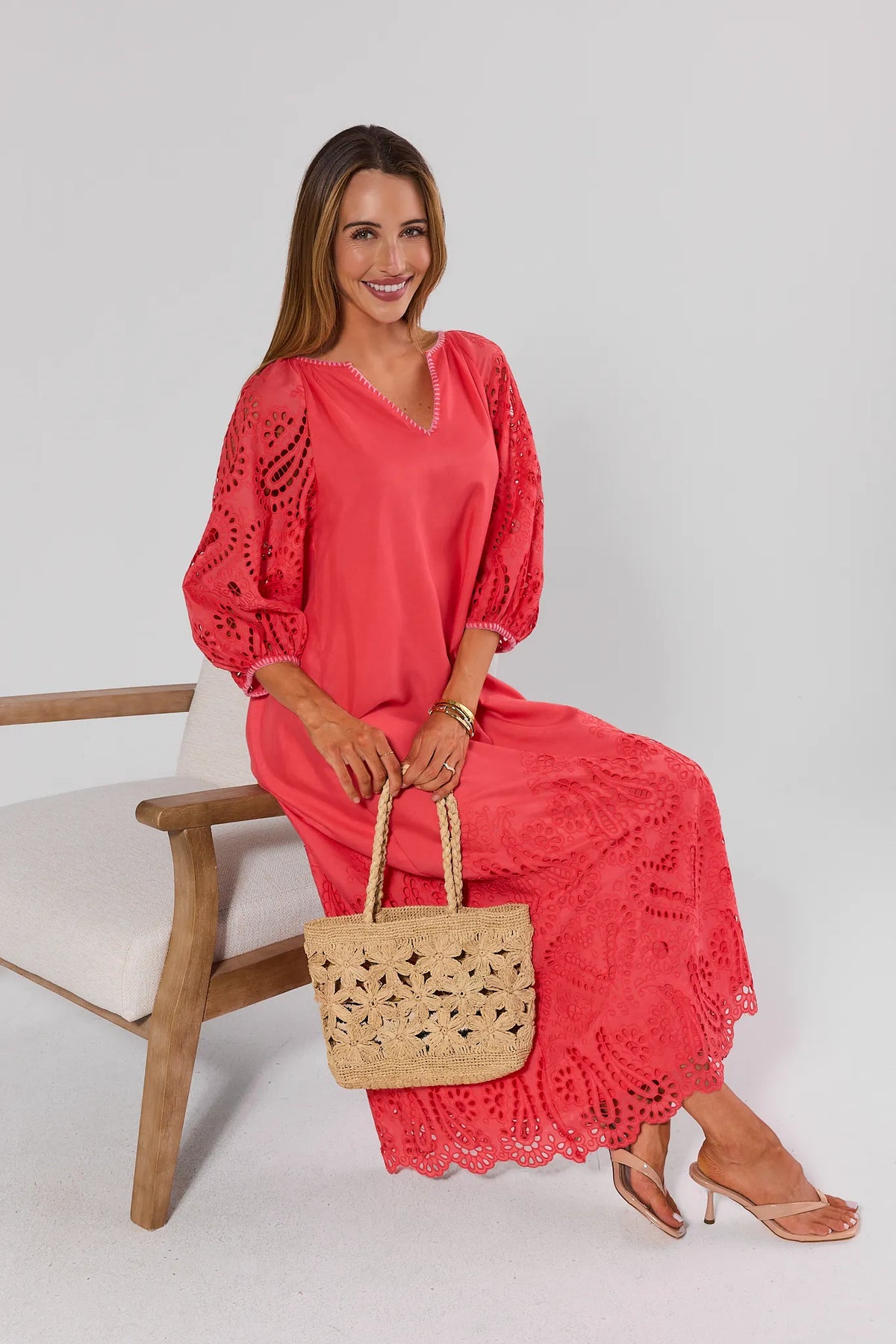 Woman seated in coral eyelet midi dress holding woven tote, showing scalloped hem detail on white chair | Avara