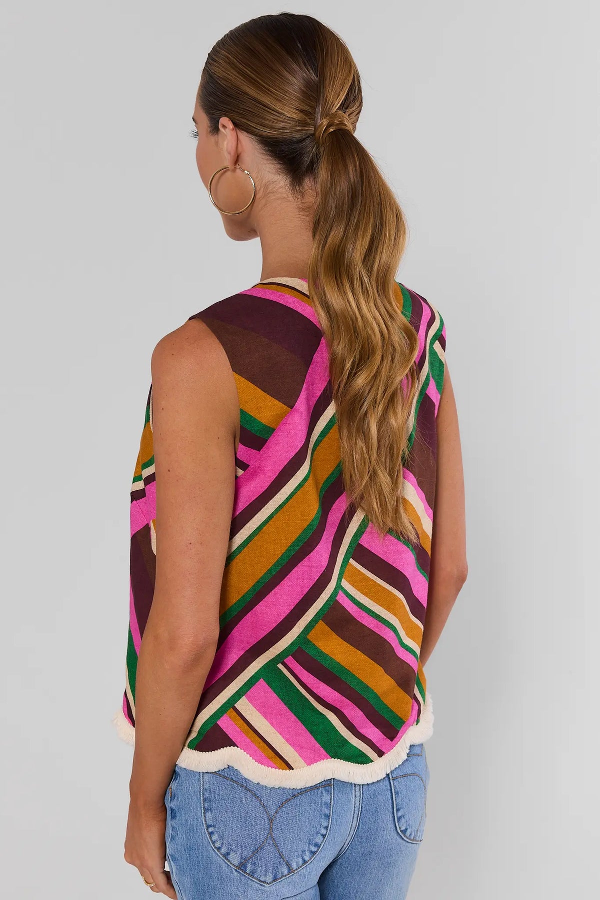 Woman turned wearing Gabby Multi Striped Fringe Trim Tank Top with jeans and gold hoop earrings on background