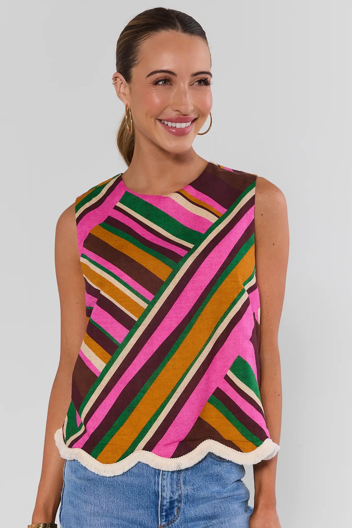 Woman wearing Gabby Multi Striped Fringe Trim Tank Top with jeans and gold hoop earrings on white background