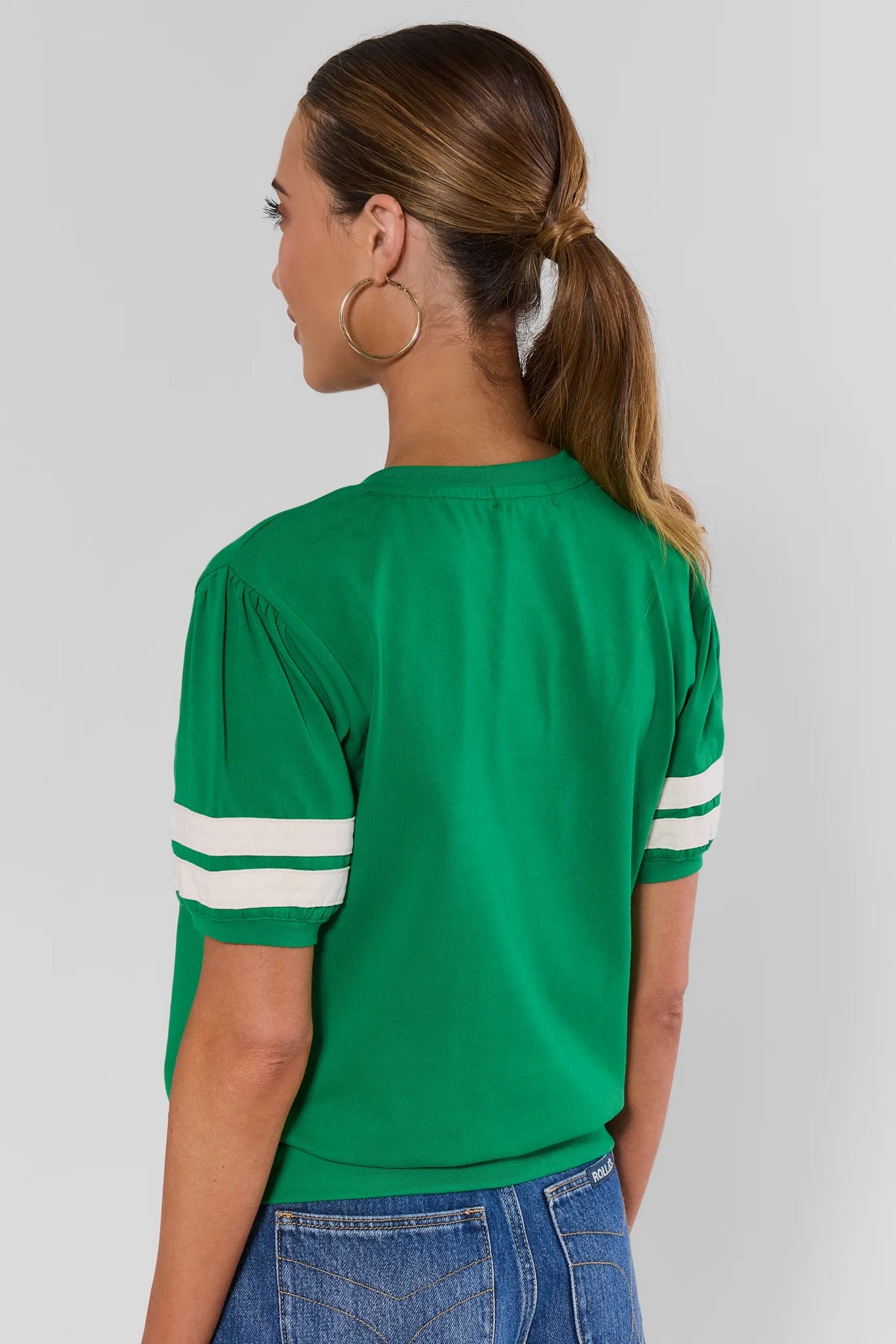 A woman shown from the back wearing a bright green short-sleeve crop top with white stripes on the sleeves, paired with blue jeans, against a light gray studio background.