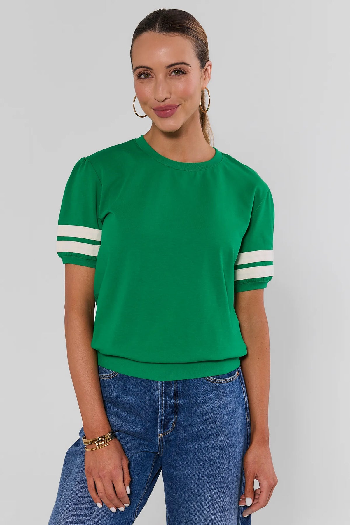 A woman modeling a bright green short-sleeve crewneck top with white stripes on the sleeves, paired with blue jeans, against a light gray studio background.