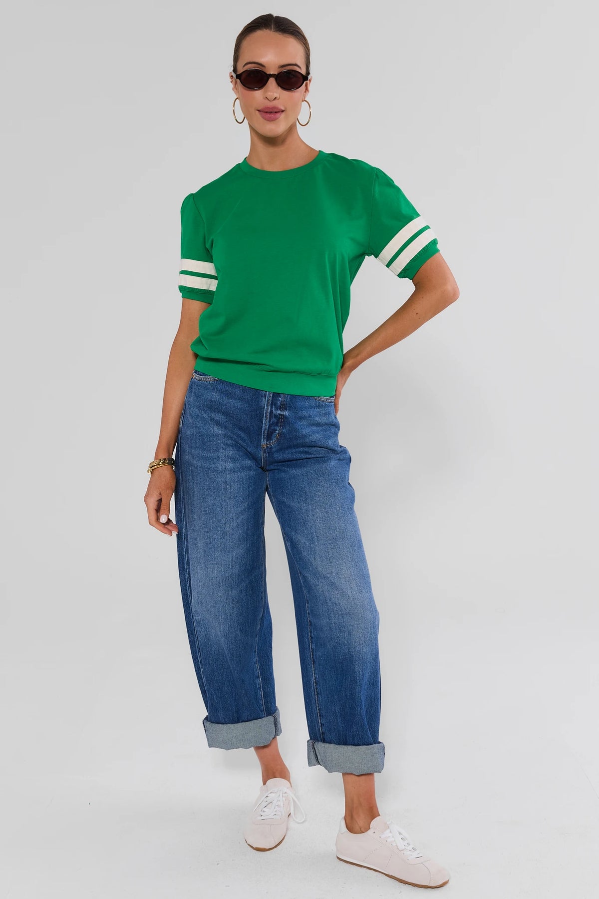 A woman modeling a bright green short-sleeve crewneck top with white stripes on the sleeves, paired with blue jeans and white sneakers, against a light gray studio background.