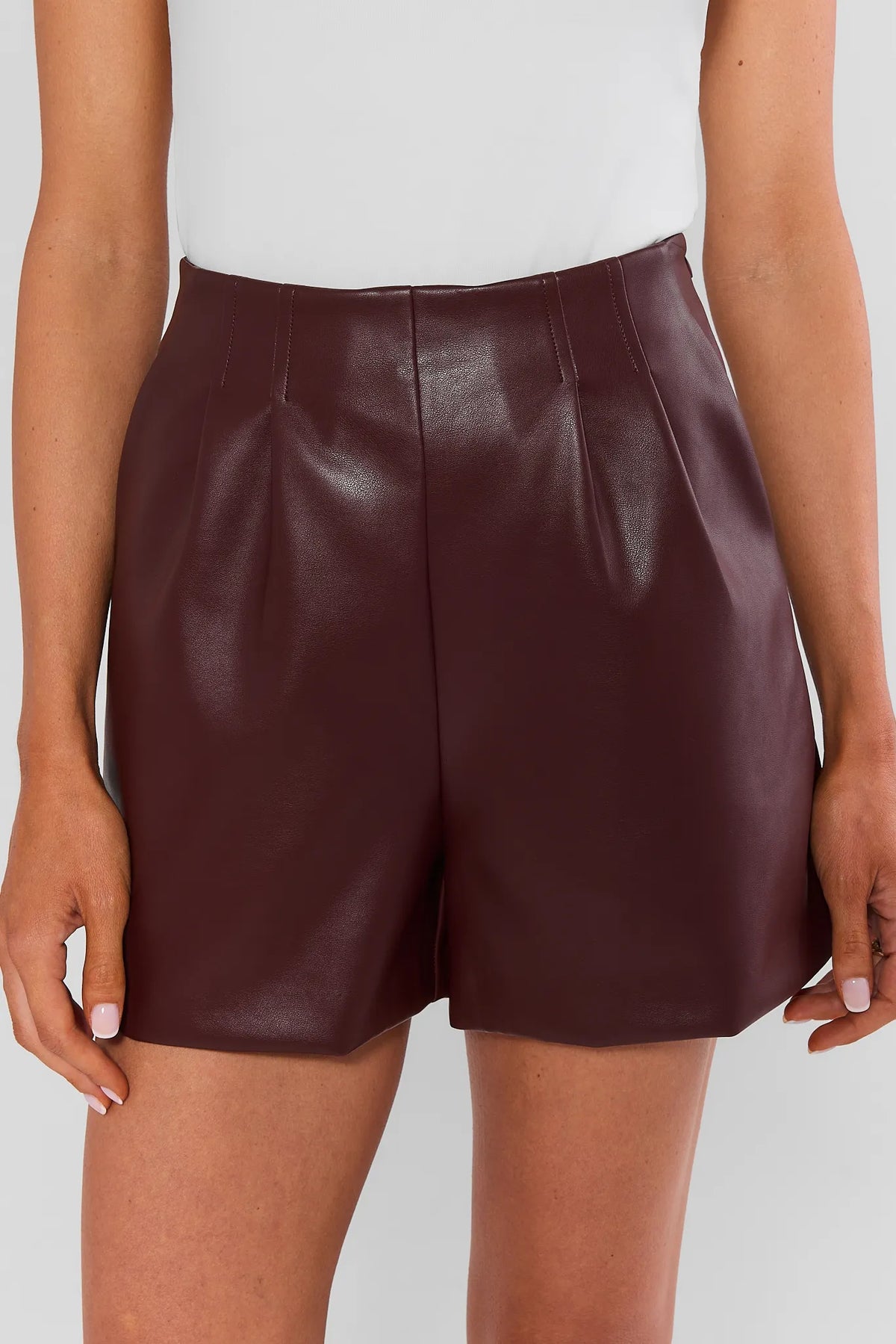 High-waisted burgundy faux leather shorts with front pleats, close-up front view on model | Avara