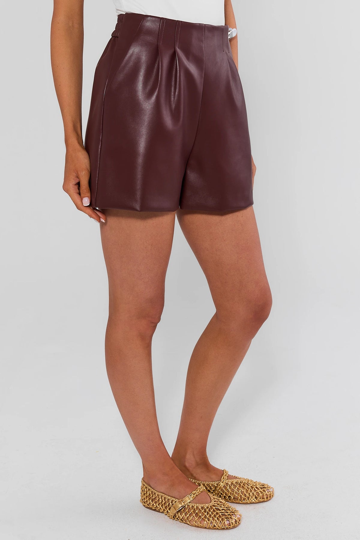 High-waisted burgundy faux leather shorts, side profile view showing pleated detail and woven flats | Avara