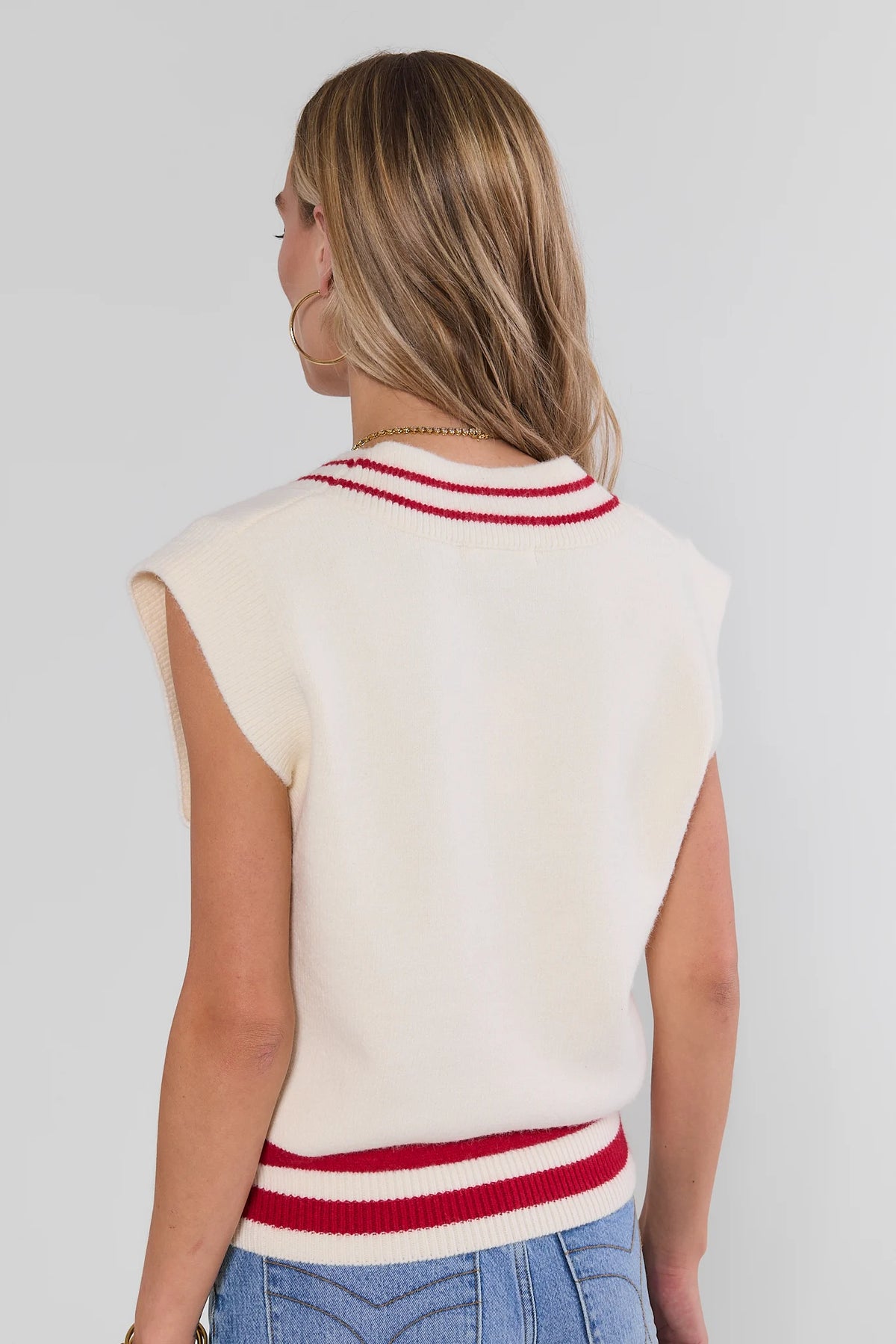 Back view of cream knit sweater vest with red contrast stripe trim at neckline and hem, paired with blue jeans | Avara