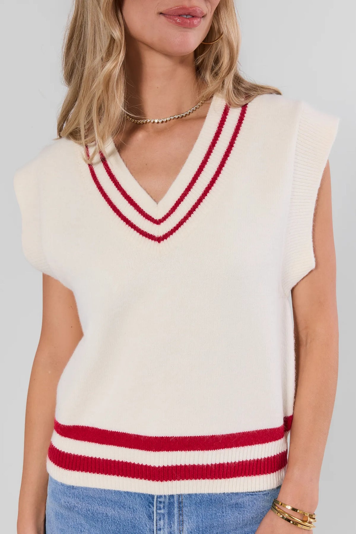 Close-up of cream knit sweater vest with red double-stripe V-neckline trim detail, worn with gold necklace | Avara