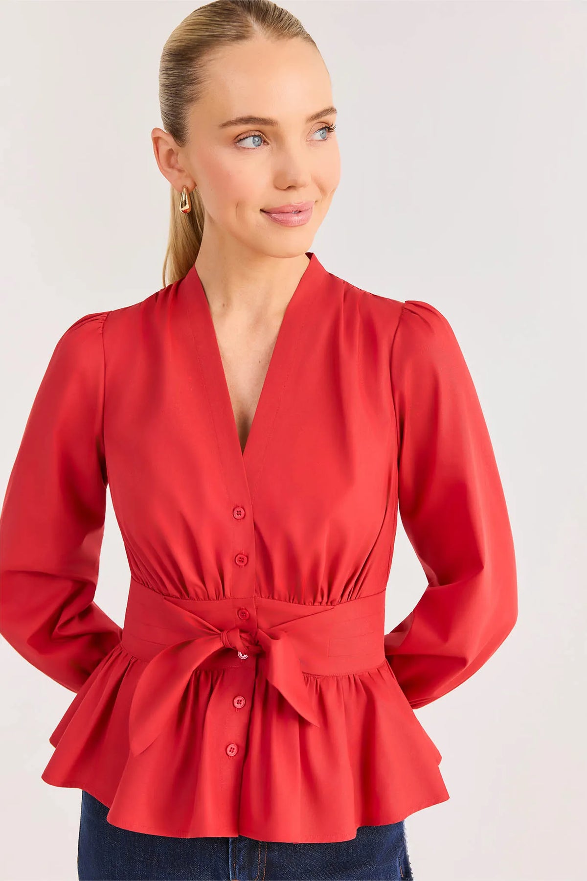 A woman wearing a red long-sleeve peplum blouse with a V-neck, button front, and matching tie belt, photographed against a light gray studio background.