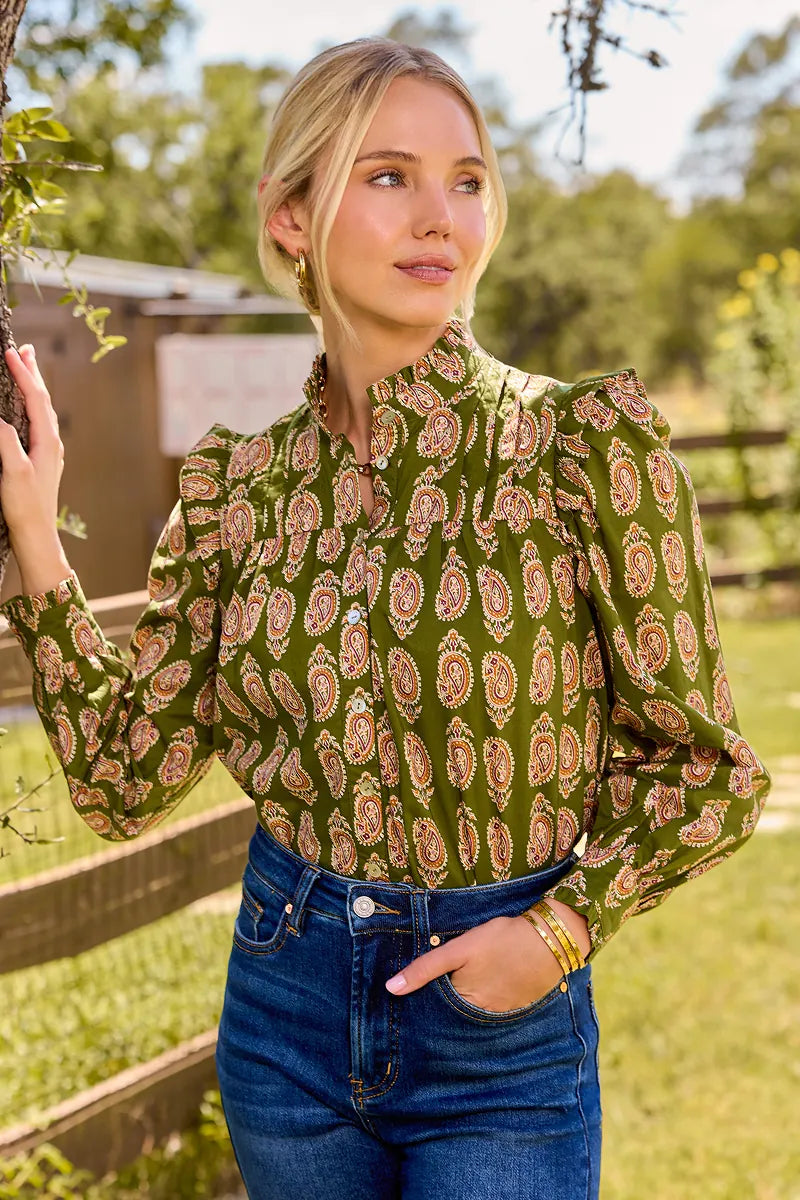 A woman wearing a green long-sleeve blouse with a brown and beige paisley pattern, paired with blue jeans, standing outdoors in front of a wooden fence and trees.