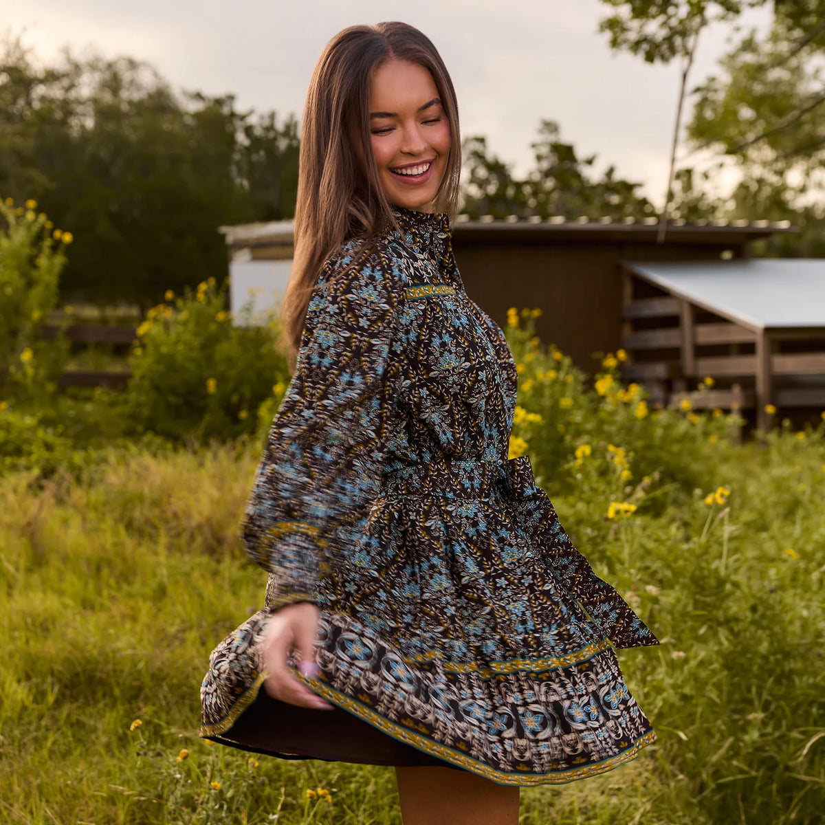 A woman outdoors in a grassy field wearing a long-sleeve, high-neck, flared dress with a blue, black, and brown bohemian-style pattern and yellow-gold trim.