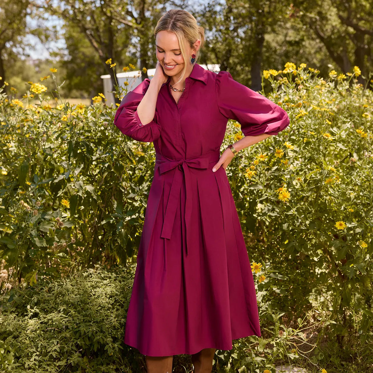 A woman wearing a long-sleeve magenta midi dress with a tie waist belt, standing outdoors in a sunlit garden with green foliage and yellow flowers.