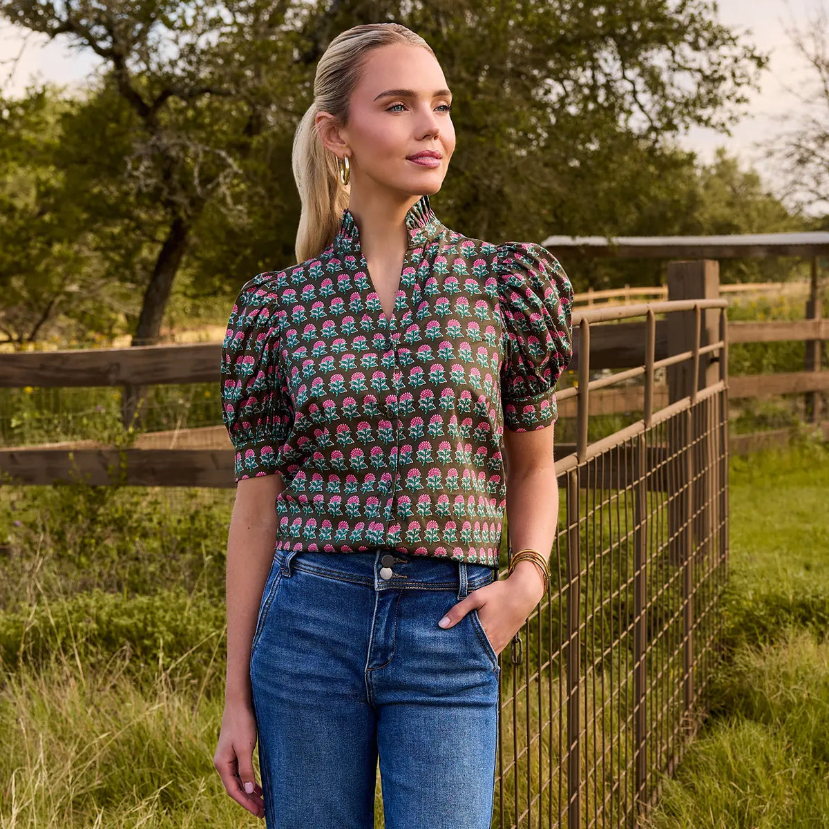 A woman stands outdoors in a grassy field beside a wooden fence, wearing a short-sleeve blouse with puff sleeves and a high collar in a green, pink, and blue paisley print, paired with blue jeans.