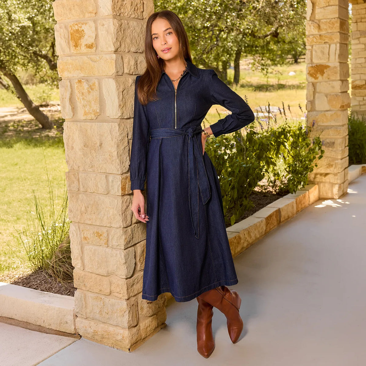 A woman wearing a dark blue denim midi dress with a zippered collar, long sleeves, and a matching tie belt, standing outdoors in front of a stone pillar.