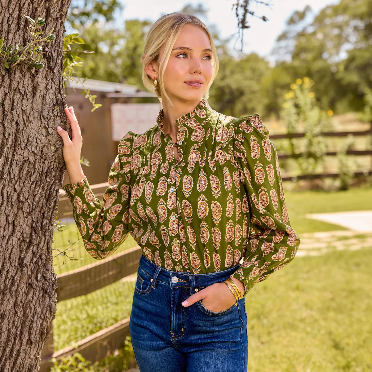 A woman wearing a green long-sleeve blouse with a brown and beige paisley pattern, standing outdoors next to a tree.