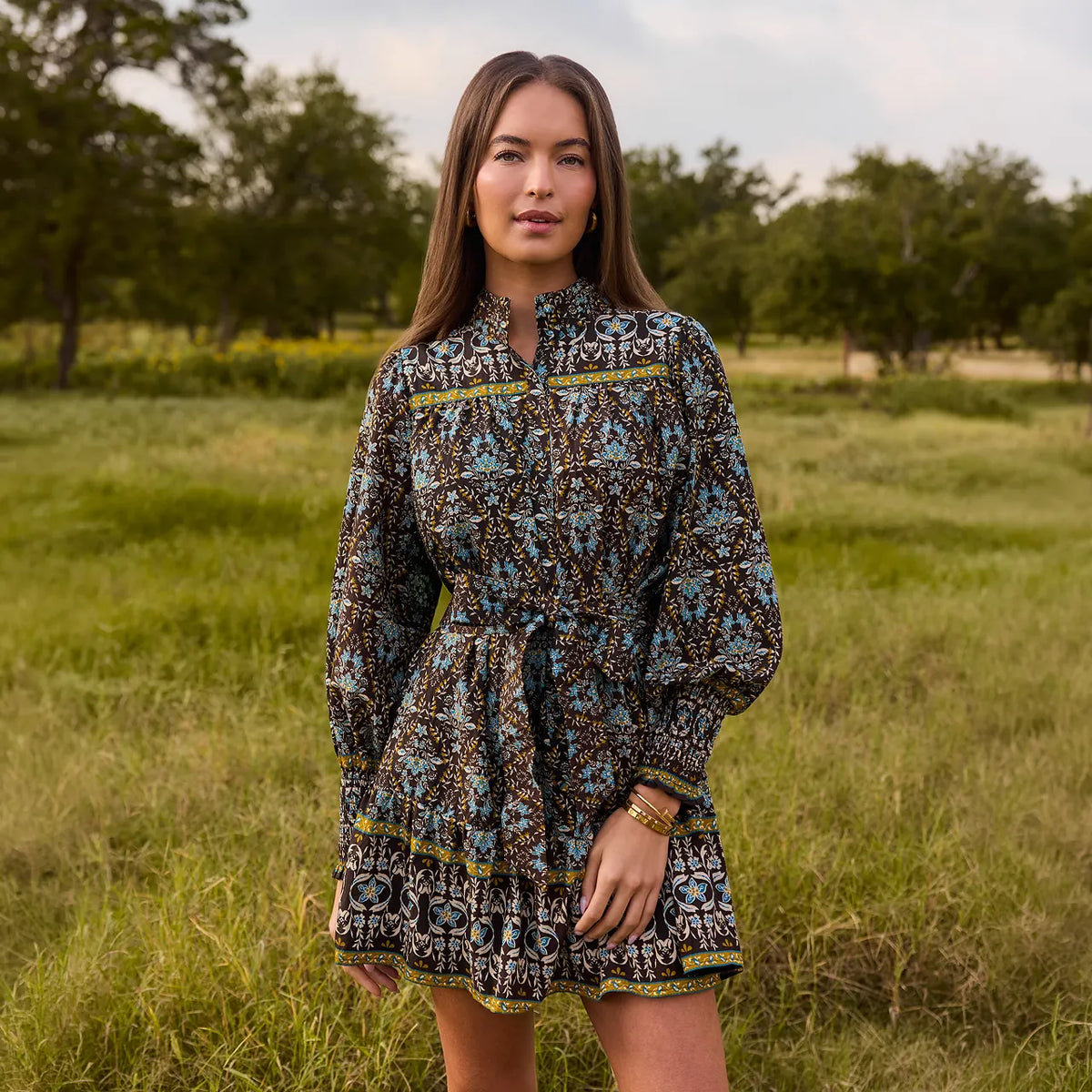 A woman stands in a grassy outdoor field wearing a long-sleeve mini dress with a detailed geometric and tribal-style pattern in black, brown, blue, and yellow.