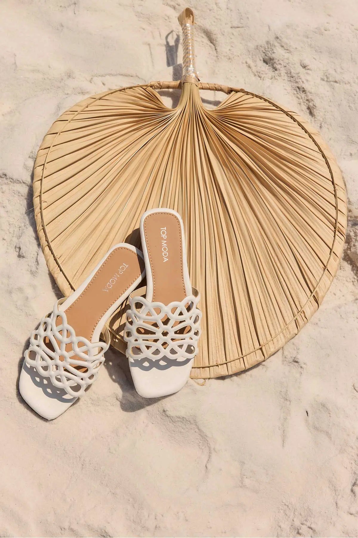 White cutout floral sandals on sandy beach surface with dried palm leaf - vacation styling | Avara