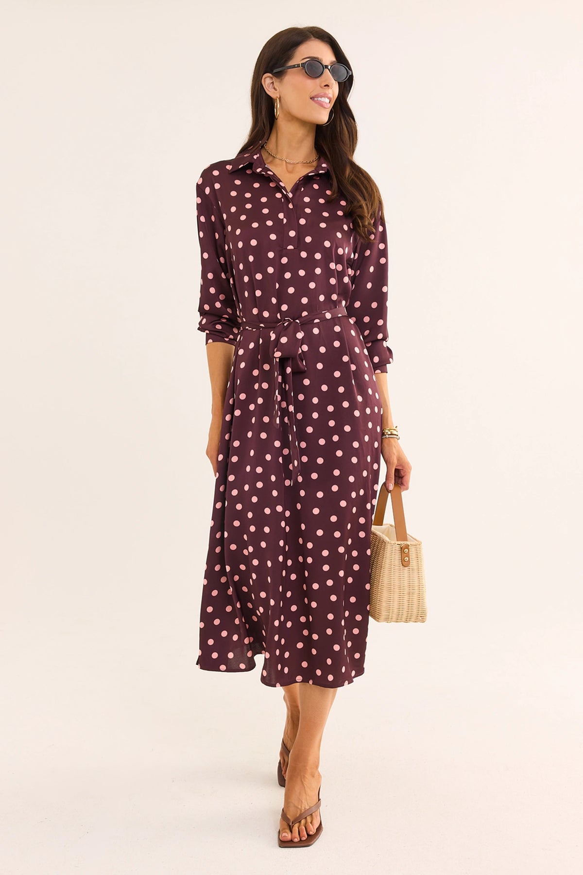 Woman walking in brown polka dot midi dress with long sleeves, tied waist, styled with sandals and wicker bag | Avara