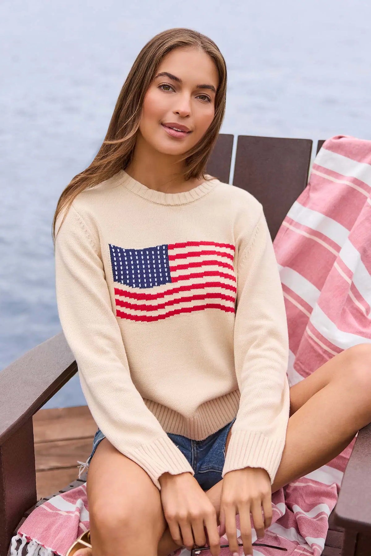 Cream American Flag Knit Sweater on model seated by water, intarsia flag detail, styled with denim shorts | Avara