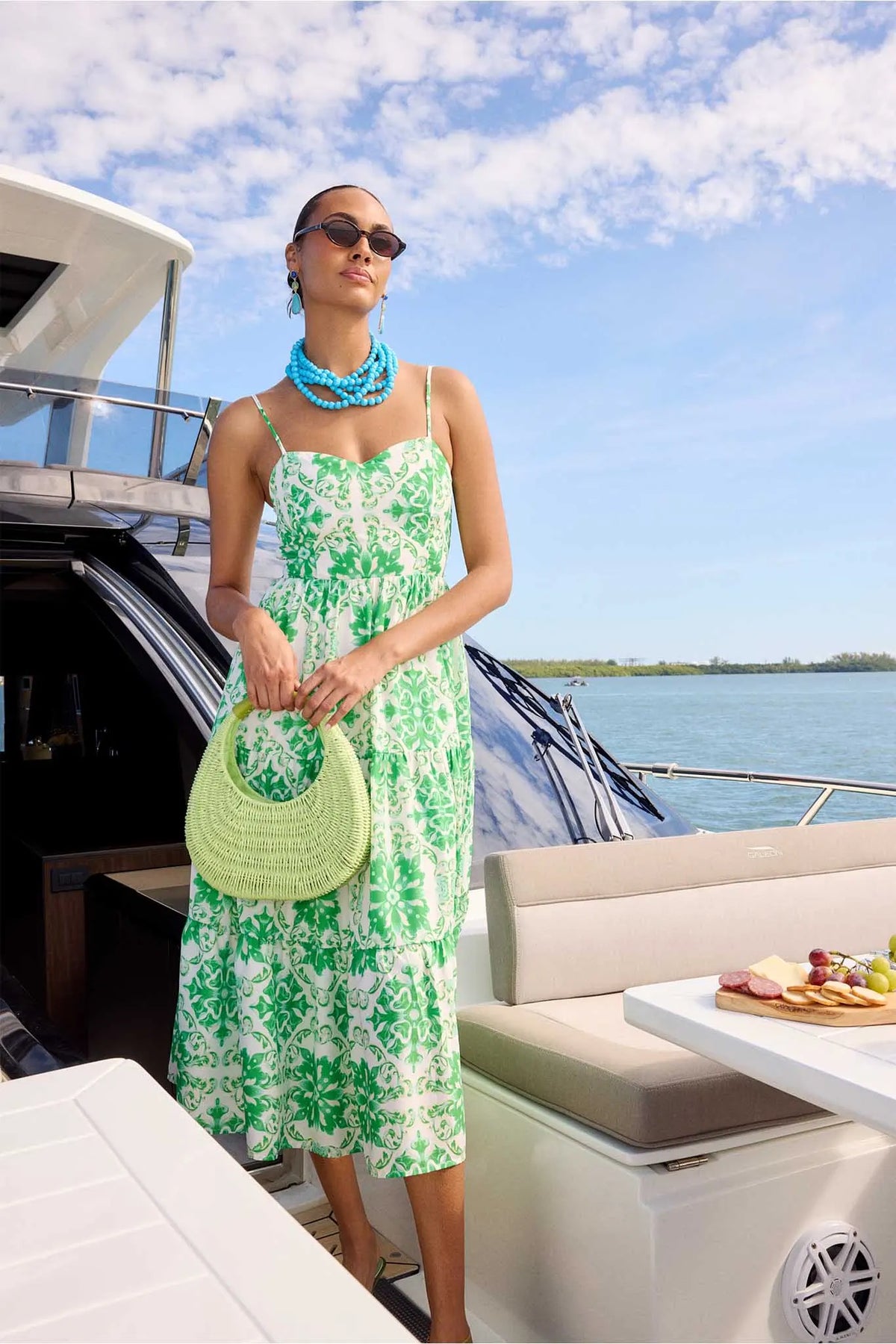 Woman on yacht wearing Lori Green Printed Midi Dress with floral pattern, chunky turquoise jewelry, and holding a pale green woven purse.