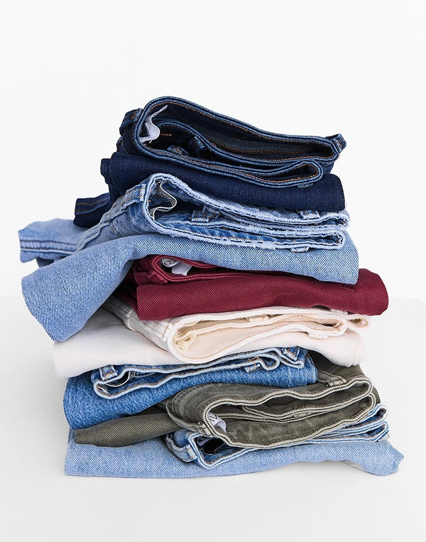 A stack of folded denim garments in various shades, including dark navy, medium blue, light blue, burgundy, cream, and olive green, photographed on a clean white background.