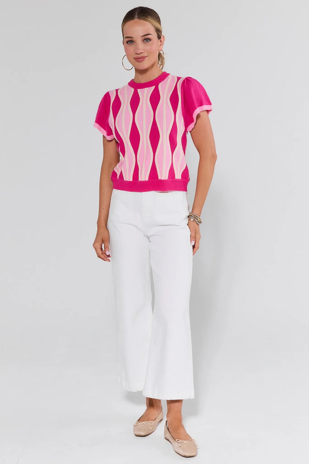 Woman wearing a pink and white patterned top with white pants on a plain background