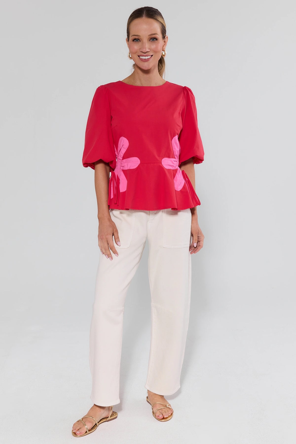 Woman wearing a red blouse with pink floral patterns and white pants on a plain background
