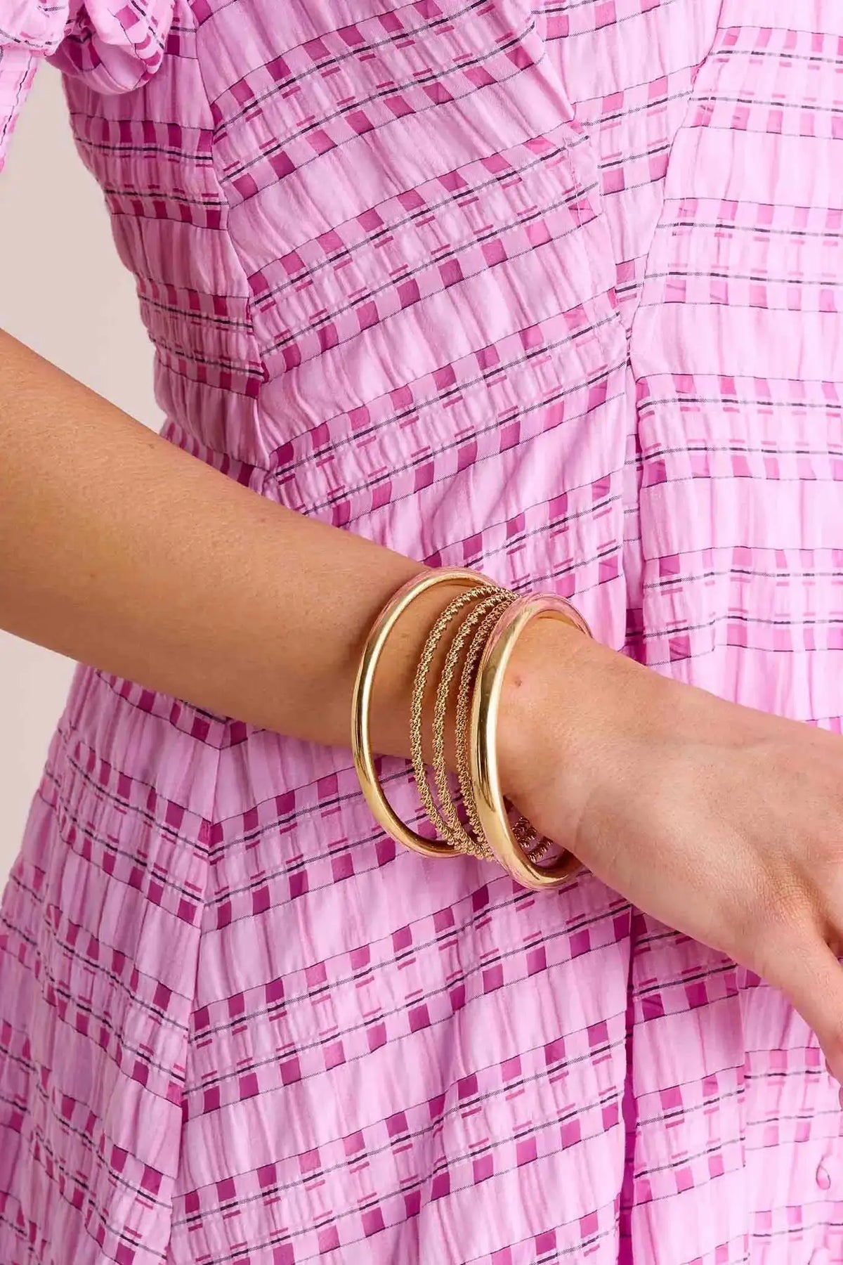 Sandra Gold Bangle Set on wrist, featuring two solid cuffs and thin rope-textured bracelets, styled against soft pink crinkled fabric.