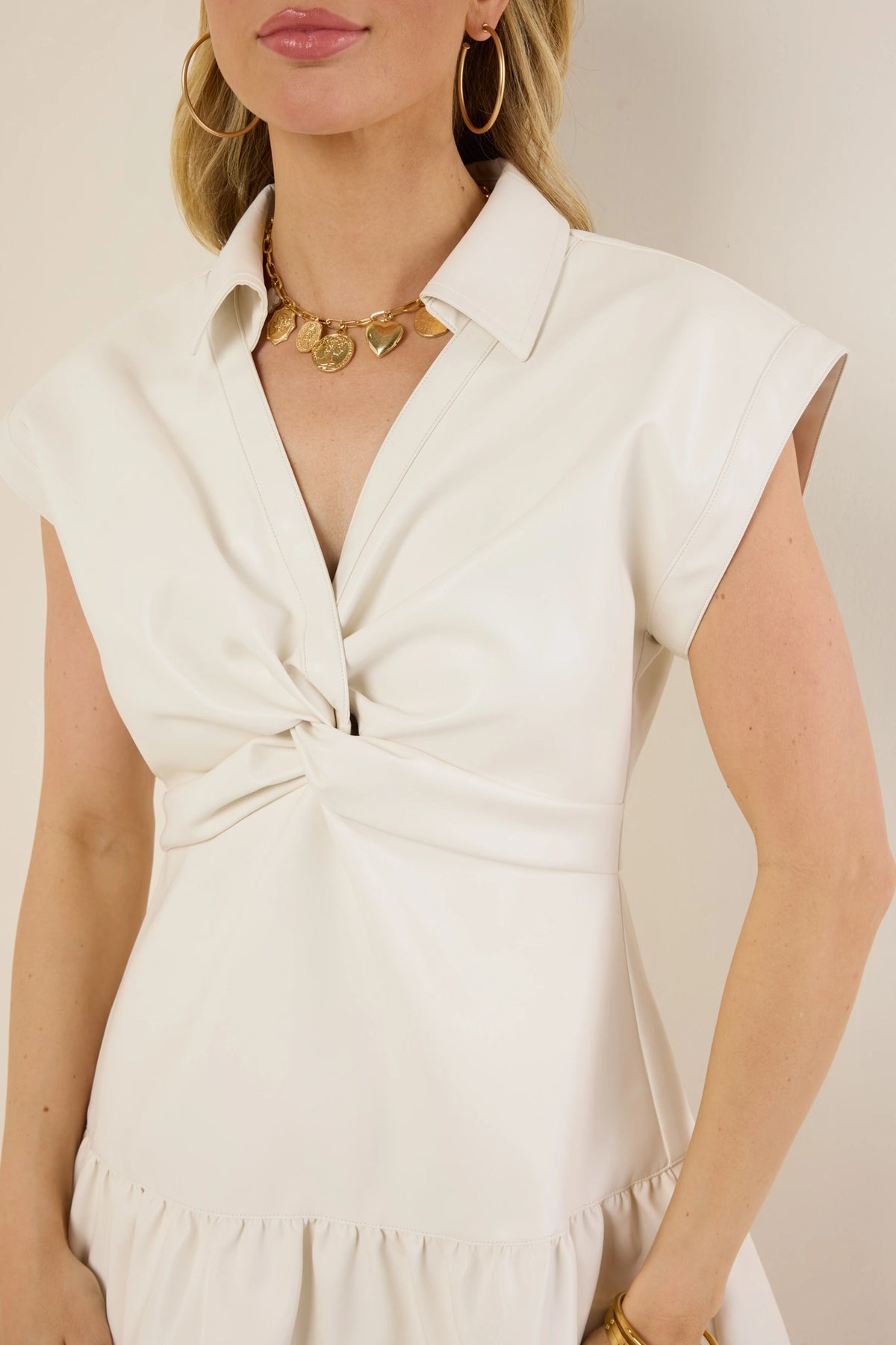Woman wearing the Sawyer Dress, a sleeveless white faux leather dress with a twist-knot bust detail, accessorized with gold jewelry.