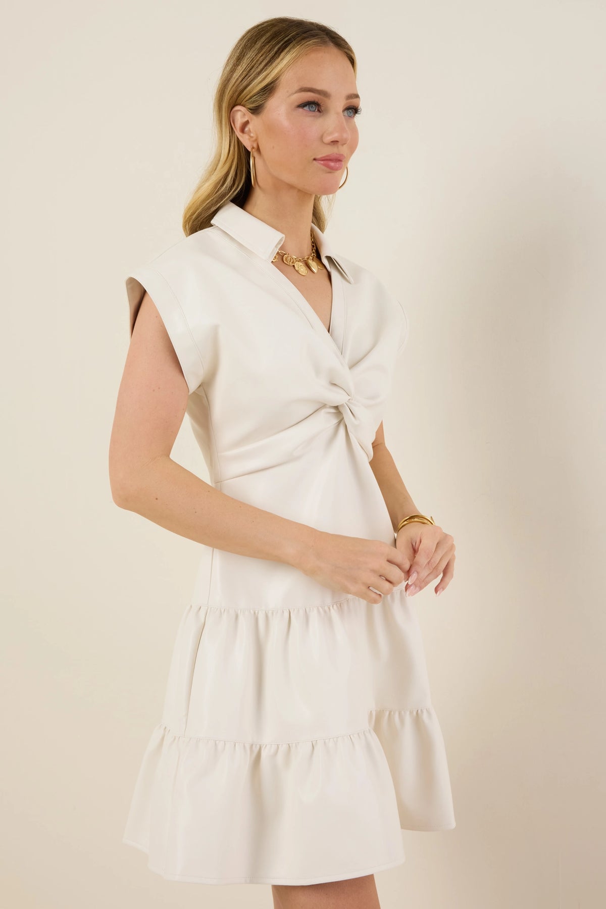 Model wearing Sawyer Dress, an ivory sleeveless faux-leather dress with a V-neck, twist detail, tiered ruffled skirt, gold accessories, and loose waves.