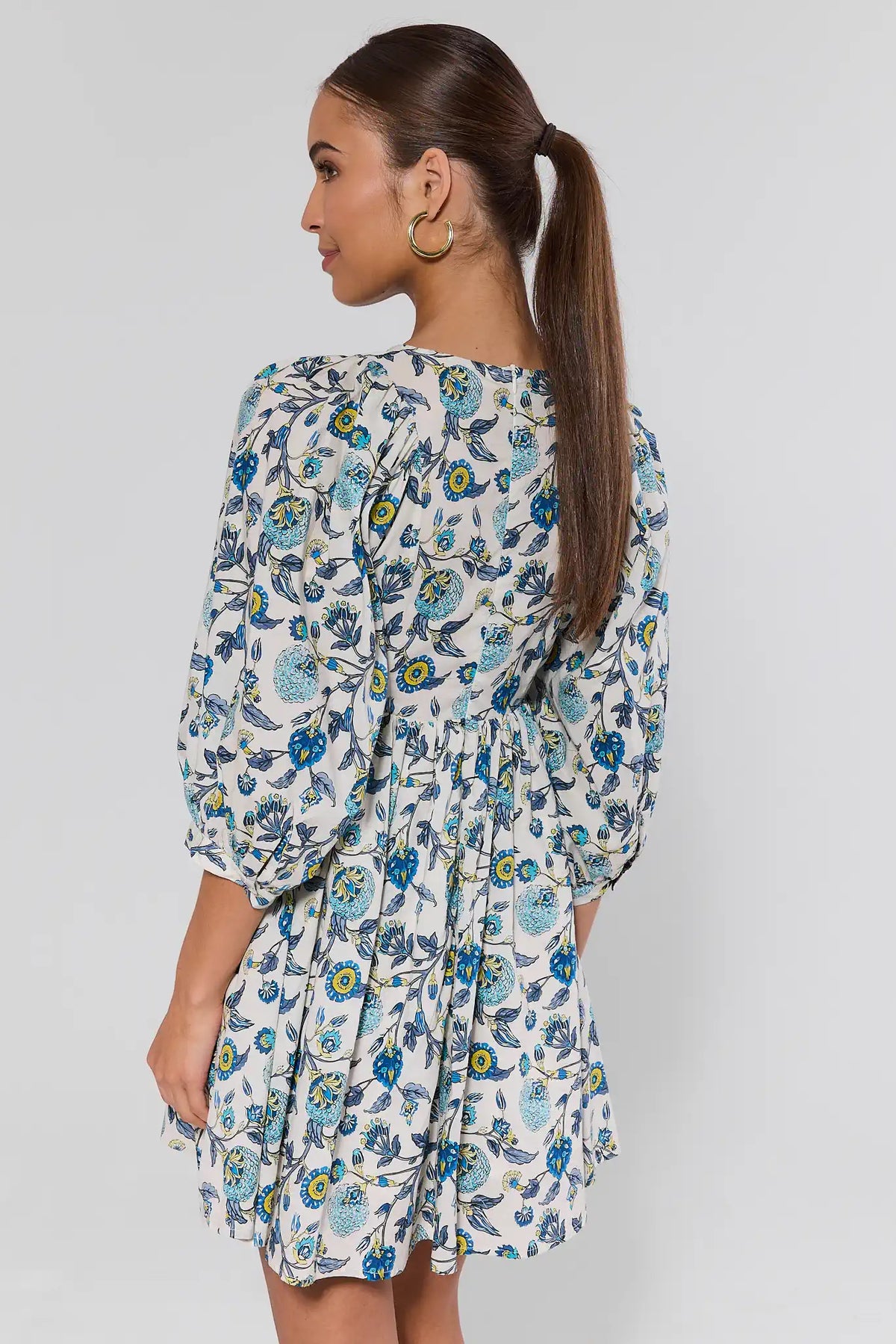 Woman in Paige Blue Floral Print Short Dress with puffed sleeves, fitted waist, flared skirt, and gold hoop earrings, viewed from the back.